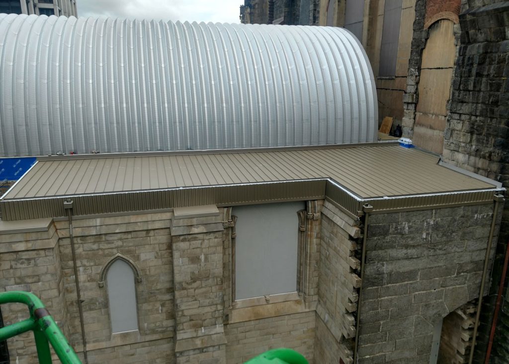 Curved silver standing seam roof above low-slope bronze metal panels on a stone building
