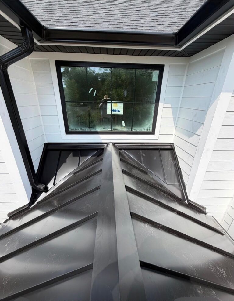 Metal Roof Repair Top-down view of a black standing seam bay roof with a center hip cap and wall flashing under a window